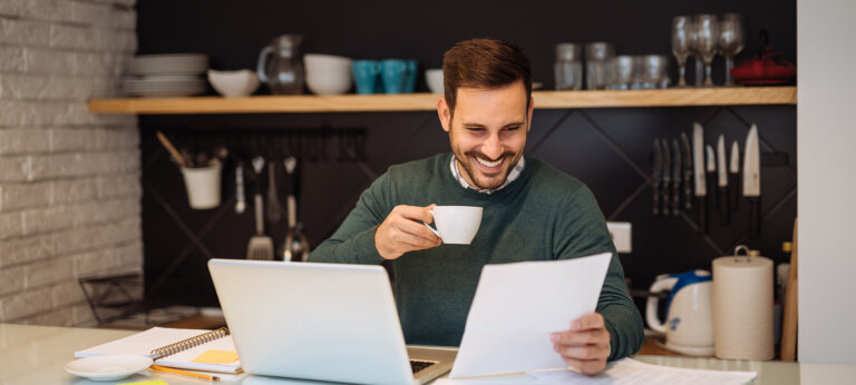 A smiling man, sitting at a laptop in a kitchen, holding a coffee and looking at an A4 piece of paper