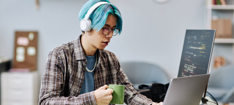 A blue-haired young man wit headphones holding a cup of coffee and staring at a laptop