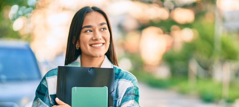 A smiling young woman looking off to the side, holding a folder and a notebook