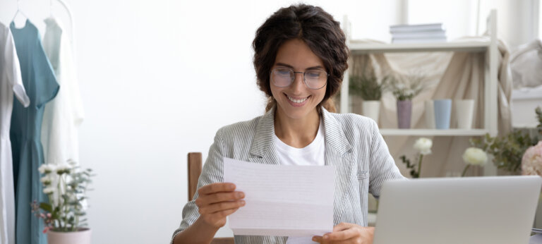 A woman working at a laptop, looking at an A4 piece of paper