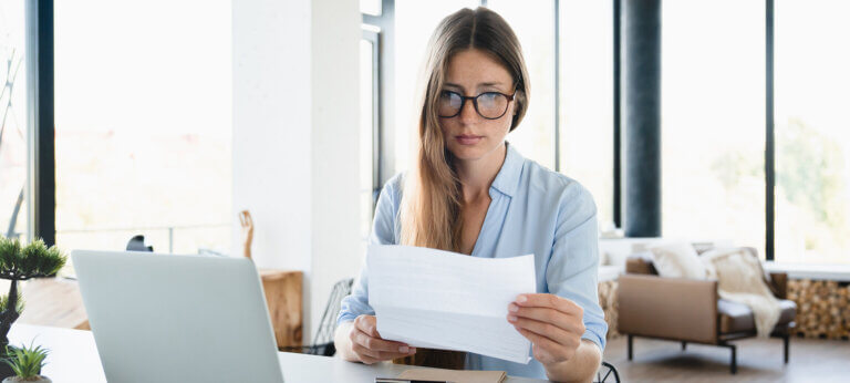 Young female employee receiving domestic bills and making payment using laptop at home desk. Caucasian freelancer woman calculating, reading report, official letter, loan mortgage