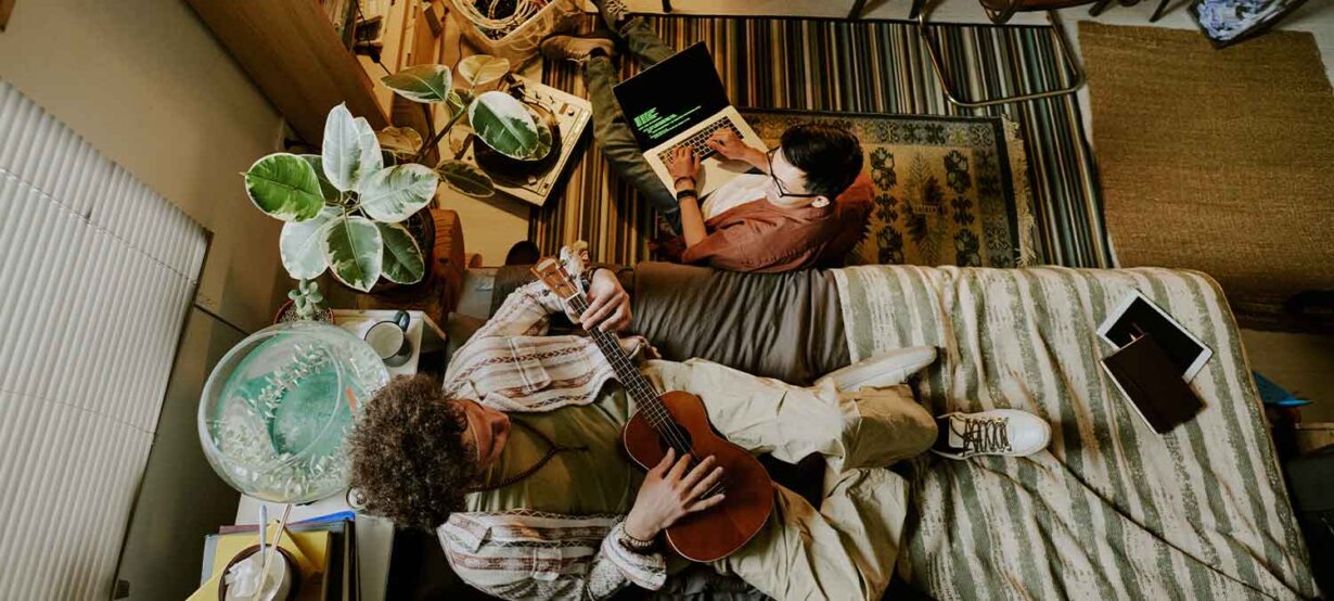A man working on his laptop while another man plays guitar
